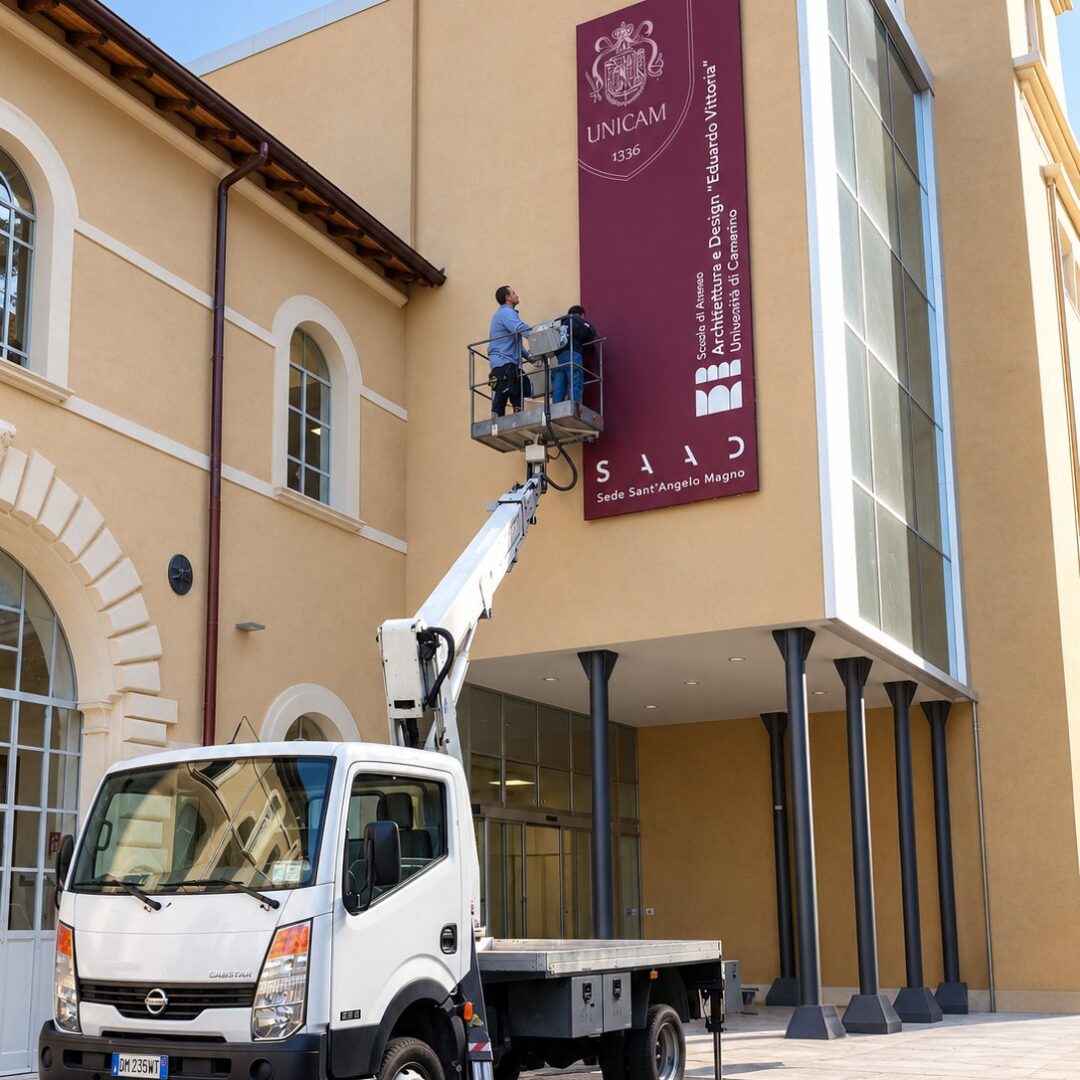 Operatore su piattaforma aerea che monta un wall banner con il logo Unicam: struttura in metallo rivestita da un telo in PVC bordeaux sulla facciata di un edificio