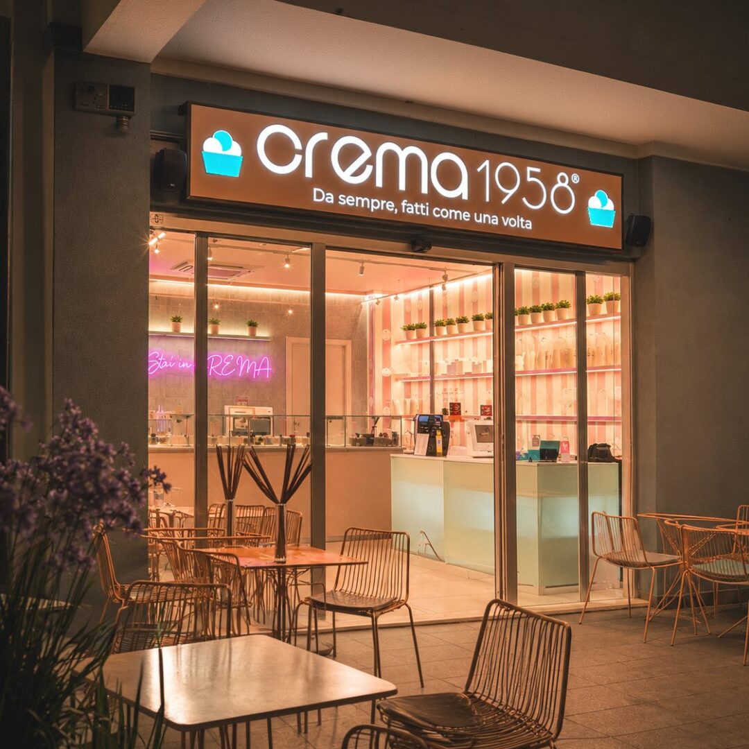 Insegna luminosa Crema 1958 sopra l'ingresso del locale, con tavolini all'aperto di sera